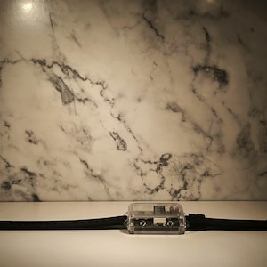 K&ouml;nnte beinhalten: Eine transparente, rechteckige Uhr mit schwarzem Armband. Das Zifferblatt ist in transparentem Kunststoff eingeschlossen und weist interne Komponenten auf. Das Armband besteht aus schwarzem Gummi und hat eine silberfarbene Schnalle. Hintergrund: wei&szlig;e Marmorwand.