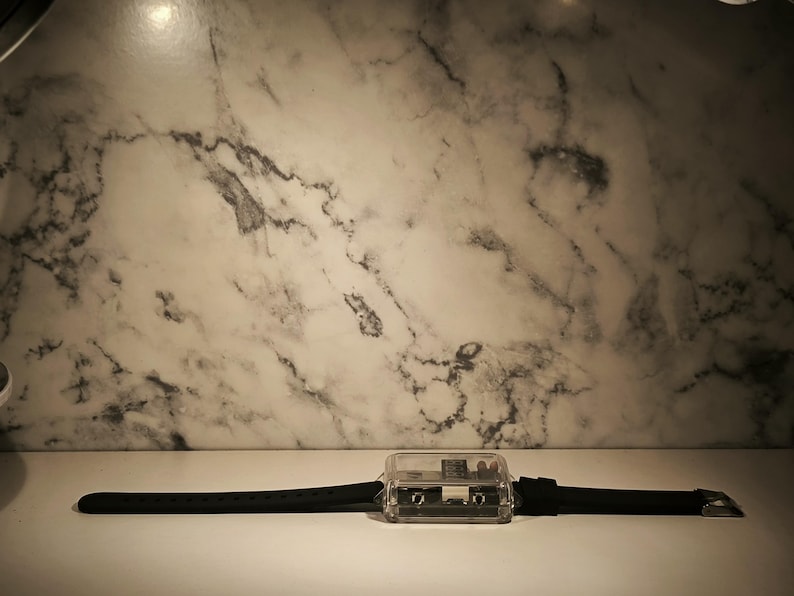 K&ouml;nnte beinhalten: Eine transparente, rechteckige Uhr mit schwarzem Armband. Das Zifferblatt ist in transparentem Kunststoff eingeschlossen und weist interne Komponenten auf. Das Armband besteht aus schwarzem Gummi und hat eine silberfarbene Schnalle. Hintergrund: wei&szlig;e Marmorwand.