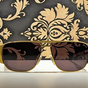 May include: Gold-framed aviator sunglasses with dark lenses. The sunglasses have a rectangular shape and a double bridge. The temples are thick and feature a textured design. The background is a black surface with a gold floral pattern.