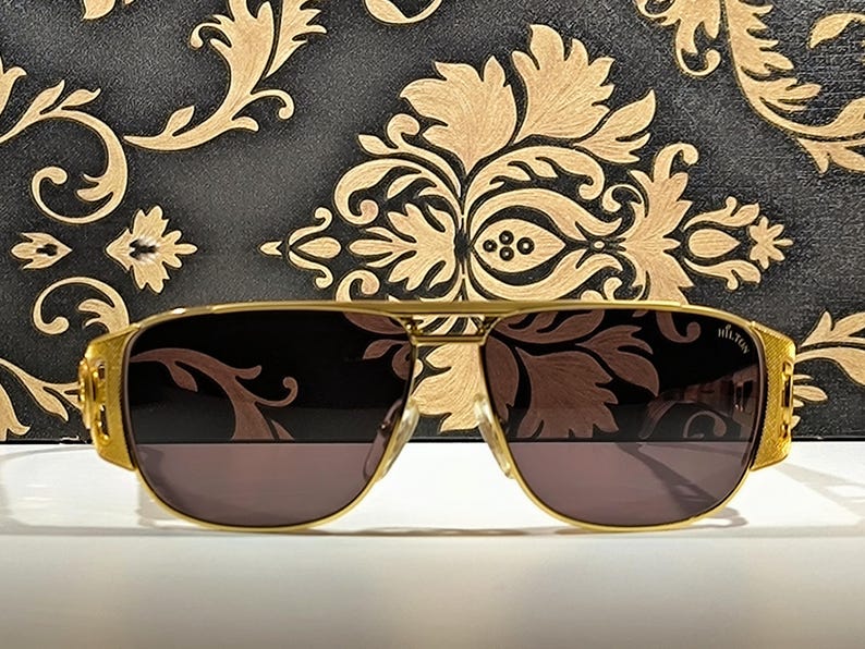 May include: Gold-framed aviator sunglasses with dark lenses. The sunglasses have a rectangular shape and a double bridge. The temples are thick and feature a textured design. The background is a black surface with a gold floral pattern.