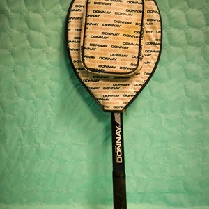 May include: A black and white tennis racket with a white and black Donnay logo on the handle. The racket head is covered in a white and black pattern with the word "Donnay" repeated.
