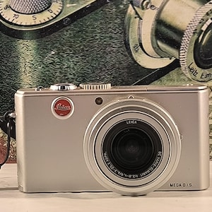 May include: A silver Leica digital camera with a black lens and a red Leica logo. The camera has a black strap and the text "MEGA O.I.S." printed on the bottom.