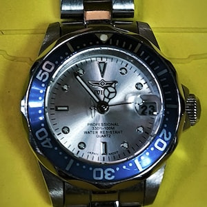 Puede incluir: Un reloj analógico de tono plateado con esfera plateada metálica, bisel giratorio azul y negro y correa de eslabones de acero inoxidable. La esfera incluye el texto Invicta, Professional, 330ft-100M, Water Resistant, Quartz y Japan Movt.