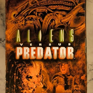 Könnte beinhalten: Eine Vintage-Videospielbox für Aliens Versus Predator. Die Box hat ein dunkelorange-braunes Farbschema mit dem Spieltitel in großen, fetten Buchstaben. Bilder des Aliens und des Predators sind ebenfalls zu sehen.