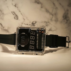 K&ouml;nnte beinhalten: Eine quadratische Digitaluhr mit transparentem Geh&auml;use und schwarzem Zifferblatt. Die Uhr hat ein schwarzes Armband und eine silberne Schnalle. Die Digitalanzeige zeigt die Uhrzeit in wei&szlig;en Ziffern an. Ein modernes Accessoire.