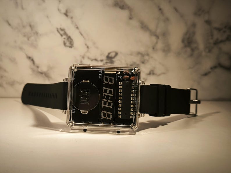 K&ouml;nnte beinhalten: Eine quadratische Digitaluhr mit transparentem Geh&auml;use und schwarzem Zifferblatt. Die Uhr hat ein schwarzes Armband und eine silberne Schnalle. Die Digitalanzeige zeigt die Uhrzeit in wei&szlig;en Ziffern an. Ein modernes Accessoire.