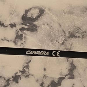 May include: Close-up of a black eyeglass arm with the white text "CARRERA CE". The arm is set against a mottled, light-colored background.