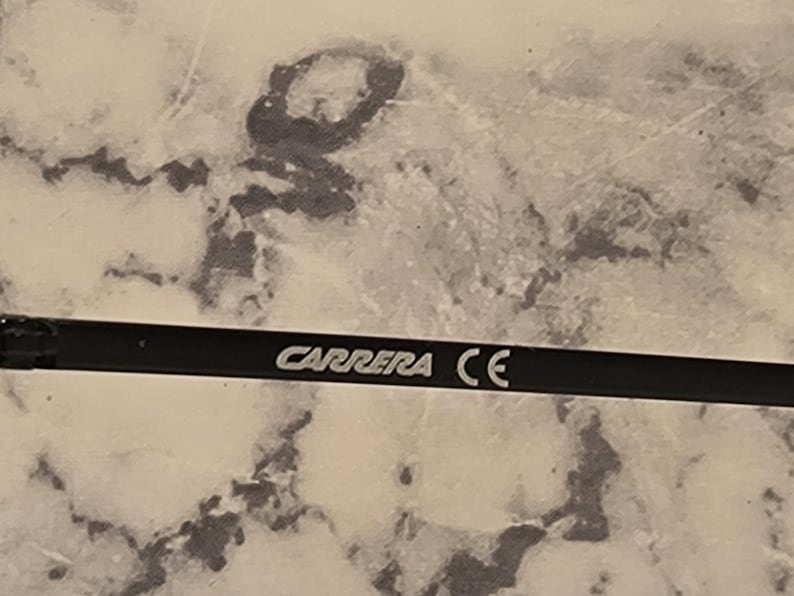 May include: Close-up of a black eyeglass arm with the white text "CARRERA CE". The arm is set against a mottled, light-colored background.
