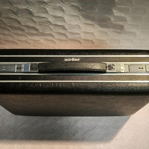 Puede incluir: Una maleta negra vintage con ribetes plateados y un asa negra. La palabra "airbus" está impresa en el asa. La maleta tiene forma rectangular y está cerrada.