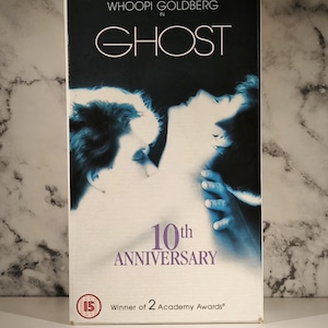 Peut inclure: Une cassette VHS du film "Ghost" avec Patrick Swayze, Demi Moore et Whoopi Goldberg. La couverture est sombre avec une image romantique de deux personnes. Le texte "10th Anniversary" est affiché, ainsi que "Winner of 2 Academy Awards".