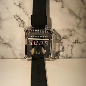 K&ouml;nnte beinhalten: Eine schwarze Digitalarmbanduhr mit einem transparenten, rechteckigen Zifferblatt. Die Uhr zeigt rote digitale Zahlen an. Das Armband ist schwarz und aus flexiblem Material. Die Uhr hat ein Retro-Futuristic-Design.
