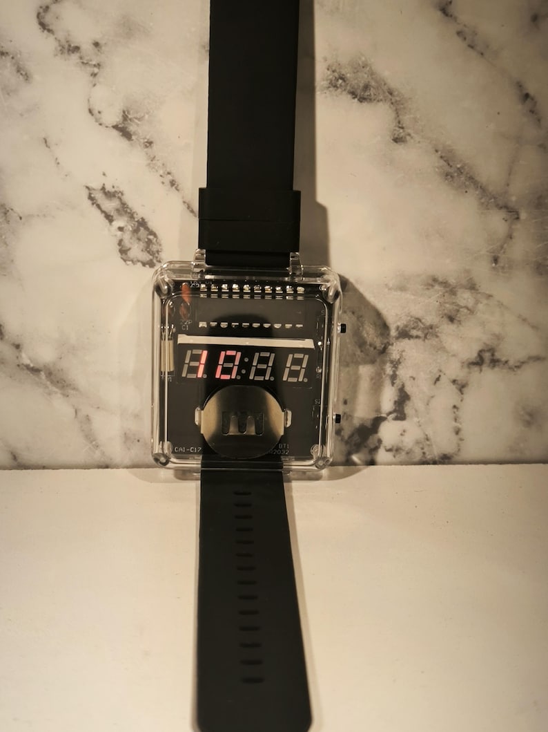 K&ouml;nnte beinhalten: Eine schwarze Digitalarmbanduhr mit einem transparenten, rechteckigen Zifferblatt. Die Uhr zeigt rote digitale Zahlen an. Das Armband ist schwarz und aus flexiblem Material. Die Uhr hat ein Retro-Futuristic-Design.