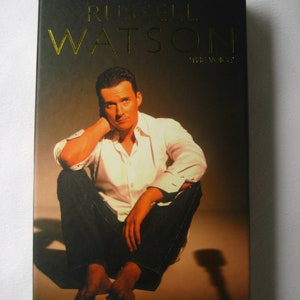 Op de afbeelding: Een DVD-hoes met een foto van Russell Watson, een zanger, zittend op de vloer met een microfoonstandaard-schaduw achter hem. De tekst op de hoes luidt "Russell Watson "The Voice" Live" en het Universal Music-logo bevindt zich rechtsonder.