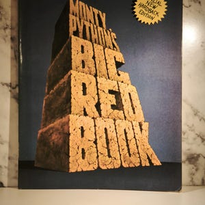 May include: The cover of "Monty Python's Big Red Book" features the title in large, textured, brick-like letters. A gold sticker indicates a special new hardback edition. The background is a gradient of dark blue to black.