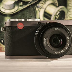 Op de afbeelding: Een zwarte en zilveren Leica digitale camera met een prominente lens. De lens is voorzien van de tekst "ELMARIT 1:2.8/24 ASPH." en "LEICA." Een rood Leica-logo is zichtbaar op de camerabody. De achtergrond toont een vage vintage camera.