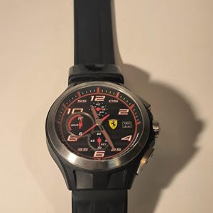 May include: Black wristwatch with a round face and a black rubber strap. The watch face features red and white detailing, with the number 12 clearly visible. The Ferrari logo is present.