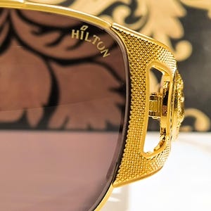 May include: Close-up of gold-colored sunglasses with a textured frame and brown lenses. The word "HILTON" is visible on the frame. The sunglasses have a decorative design on the side.