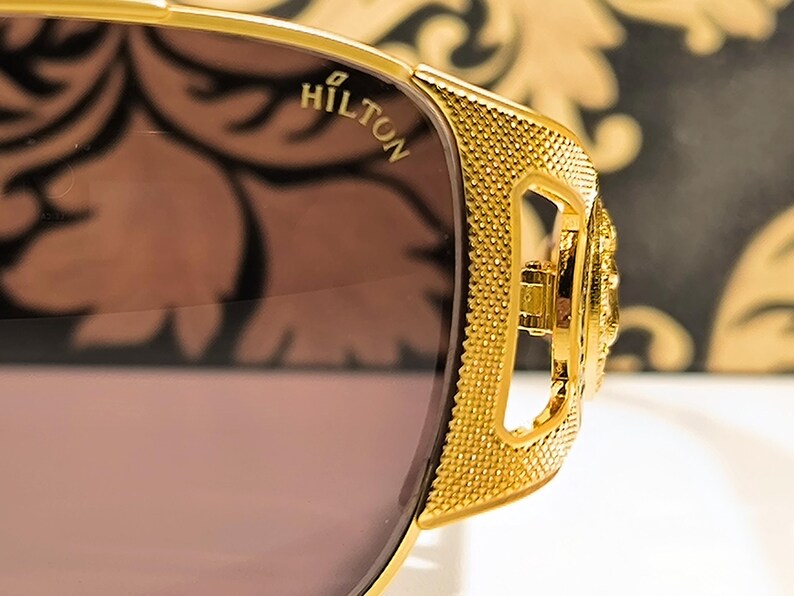 May include: Close-up of gold-colored sunglasses with a textured frame and brown lenses. The word "HILTON" is visible on the frame. The sunglasses have a decorative design on the side.