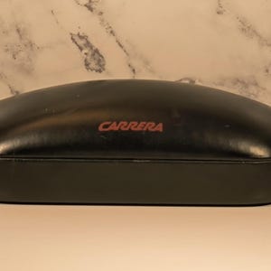 May include: A black, oval-shaped Carrera eyeglasses case. The case has a smooth, glossy finish and a red "CARRERA" logo on the top. The case is closed and appears to be made of a durable material, designed to protect eyewear.