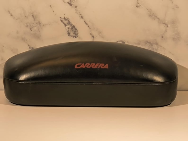 May include: A black, oval-shaped Carrera eyeglasses case. The case has a smooth, glossy finish and a red "CARRERA" logo on the top. The case is closed and appears to be made of a durable material, designed to protect eyewear.