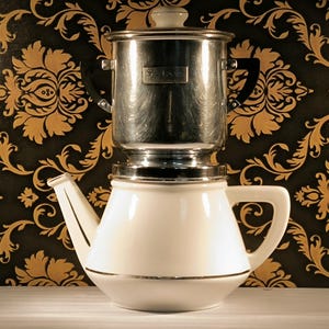 Op de afbeelding: Een witte keramische koffiepot met een zilveren bovenkant en een zilveren filter. De koffiepot heeft een gouden rand rond de basis en een gouden handvat. De koffiepot staat op een zwart-gouden gepatroneerd oppervlak.