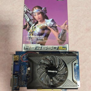 May include: A GIGABYTE HD 4850 1GB GDDR2 graphics card with a black fan and a blue circuit board. A purple user manual with a woman holding a futuristic weapon is behind the card. The manual has the text "GIGABYTE" and "ATI Radeon".
