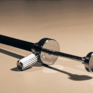Può includere: Primo piano di un rilascio cavo per fotocamera color argento con un'impugnatura nera strutturata. Il rilascio ha una piccola manopola zigrinata e un pulsante circolare. La parola "JAPAN" è visibile sul pulsante circolare.