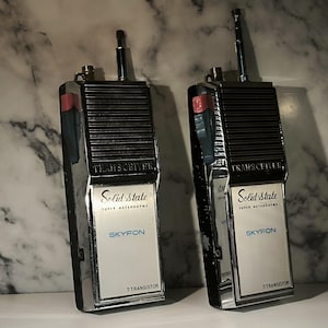 May include: Two vintage Skyfon 7 Transistor Solid State Super Heterodyne transceivers. Each handheld device features a black speaker grille, a silver faceplate with blue text, a retractable metal antenna, and a red side-mounted push-to-talk button.