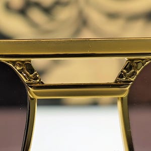 May include: Close-up of gold-colored sunglasses with a decorative bridge and dark lenses. The frame has intricate detailing near the hinges, suggesting a luxury design. The lenses are a gradient brown color.