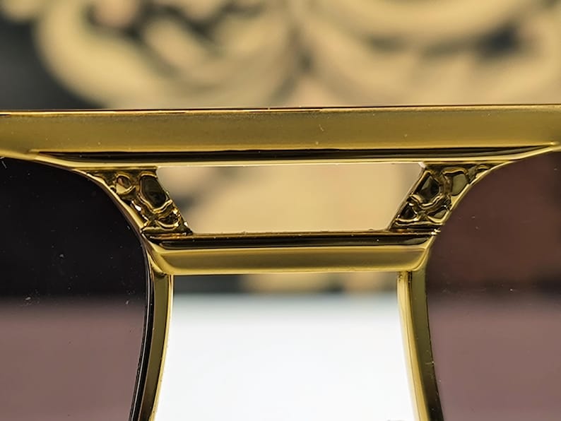 May include: Close-up of gold-colored sunglasses with a decorative bridge and dark lenses. The frame has intricate detailing near the hinges, suggesting a luxury design. The lenses are a gradient brown color.