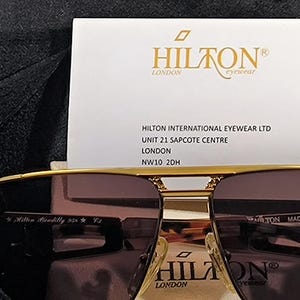 May include: Gold-framed sunglasses with brown lenses, featuring the "HILTON LONDON" logo on the frame and a white card with the brand's name and address. The sunglasses are marked "Hilton Piccadilly 958 C3".