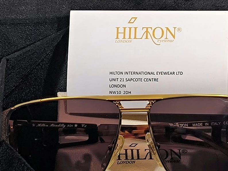 May include: Gold-framed sunglasses with brown lenses, featuring the "HILTON LONDON" logo on the frame and a white card with the brand's name and address. The sunglasses are marked "Hilton Piccadilly 958 C3".