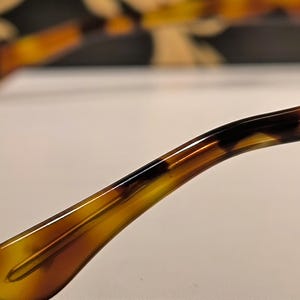 May include: Close-up of the temple arm of eyeglasses with a tortoiseshell pattern. The arm curves gently, transitioning from a light amber to a darker brown and black. The background is blurred, suggesting a pair of glasses.