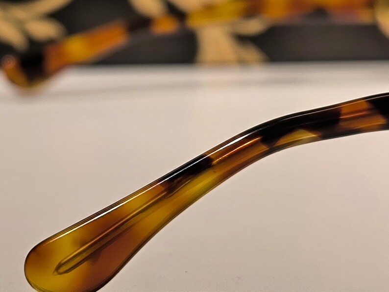 May include: Close-up of the temple arm of eyeglasses with a tortoiseshell pattern. The arm curves gently, transitioning from a light amber to a darker brown and black. The background is blurred, suggesting a pair of glasses.
