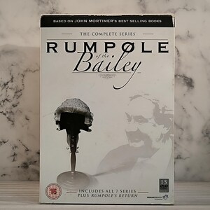 Pode incluir: Um conjunto de DVD da série completa da série de televisão britânica "Rumpole of the Bailey". O conjunto de caixas apresenta uma imagem em preto e branco de um homem com uma peruca e uma imagem em preto e branco de uma peruca de advogado em um suporte. O texto na caixa diz "The Complete Series" e "Includes all 7 series plus Rumpole's Return".