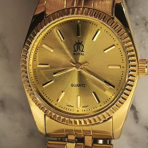 May include: A gold-toned Royal Quartz watch with a textured dial and a matching gold band. The watch face features gold hands, hour markers, and the word "QUARTZ". The watch has a fluted bezel and a crown on the right side.
