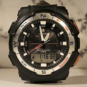 May include: Black Casio Twin Sensor watch with a silver bezel and orange accents. The watch face displays the time, compass directions, and the text "WATER RESIST 100M". The digital display shows "1:16 32".