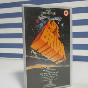 Op de afbeelding: Een VHS-cassettehoes voor de film "Monty Python's Life of Brian". De hoes is zwart met een afbeelding van de titelkaart van de film aan de voorkant. De titelkaart is een gele en oranje grafische weergave van de titel van de film, "Life of Brian", met een ruimteschip dat in de achtergrond vliegt. De hoes bevat ook de leeftijdsclassificatie van de film, "15", en de cast en crew van de film.