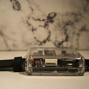 K&ouml;nnte beinhalten: Eine transparente, rechteckige Uhr mit schwarzem Armband. Das Zifferblatt ist durch das transparente Geh&auml;use sichtbar und zeigt die inneren Komponenten. Das Armband besteht aus flexiblem, schwarzem Material.
