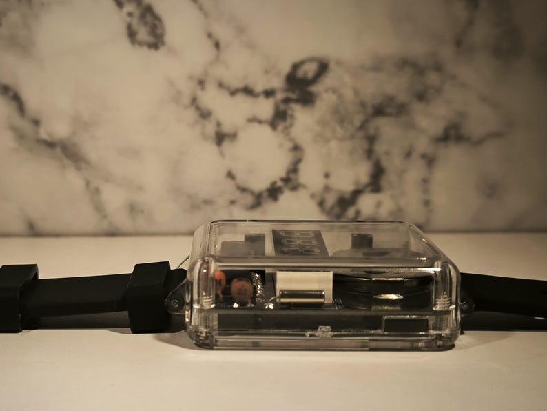 K&ouml;nnte beinhalten: Eine transparente, rechteckige Uhr mit schwarzem Armband. Das Zifferblatt ist durch das transparente Geh&auml;use sichtbar und zeigt die inneren Komponenten. Das Armband besteht aus flexiblem, schwarzem Material.