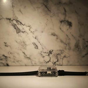 K&ouml;nnte beinhalten: Eine schwarze Armbanduhr mit einem transparenten, rechteckigen Zifferblatt, das einer Kassette &auml;hnelt. Das schwarze Armband besteht aus flexiblem Material. Der Hintergrund ist eine marmormusterte Wand.