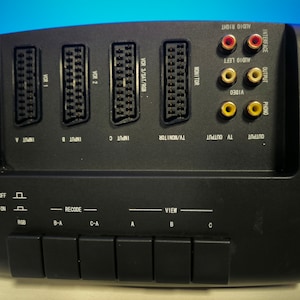 Könnte beinhalten: Schwarzer AV-Umschalter mit drei RCA-Eingängen und -Ausgängen, drei SCART-Eingängen und einem Monitorausgang. Der Umschalter verfügt über Tasten zum Auswählen von Ein- und Ausgang sowie einen Schalter zum Ein- und Ausschalten des Geräts.