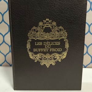 May include: A black leather-bound book with gold lettering on the cover. The title is "Les Delices du Buffet Froid".