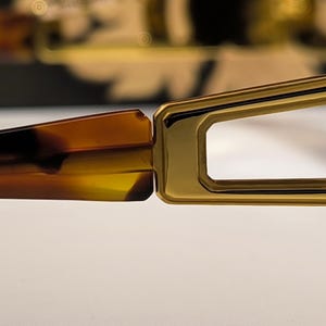 May include: Close-up of eyeglasses with a gold-colored frame and tortoise-shell arms. The rectangular lenses are visible, and the frame has a geometric design. The glasses are set against a neutral background.