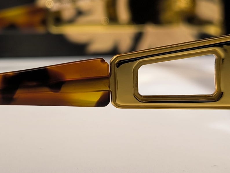 May include: Close-up of eyeglasses with a gold-colored frame and tortoise-shell arms. The rectangular lenses are visible, and the frame has a geometric design. The glasses are set against a neutral background.
