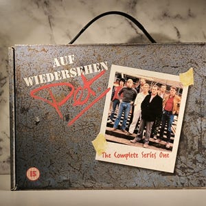 Pode incluir: Um box set com uma alça, apresentando o texto "AUF WIEDERSEHEN PET" em branco e vermelho, e uma foto de um grupo de pessoas. A foto está rotulada como "The Complete Series One". A caixa tem uma aparência metálica desgastada.
