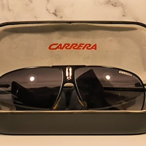 May include: Black Carrera sunglasses with dark lenses rest in an open black case. The brand name "CARRERA" is printed in red on the case and on the sunglasses. The case has a light gray interior.