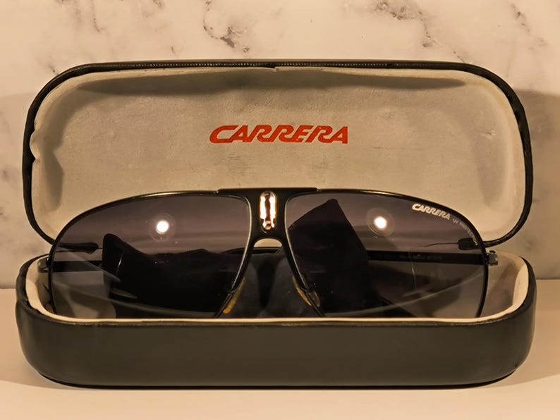 May include: Black Carrera sunglasses with dark lenses rest in an open black case. The brand name "CARRERA" is printed in red on the case and on the sunglasses. The case has a light gray interior.