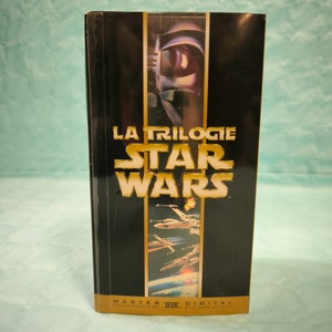 May include: A black VHS tape case with gold lettering that reads "La Trilogie Star Wars" and "Master THX Digital Pour une qualite de son et d'image optimale". The case features a black and white image of Darth Vader's helmet and a scene from the movie with X-Wing fighters.