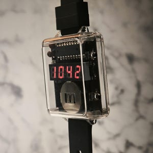 K&ouml;nnte beinhalten: Eine digitale Armbanduhr mit einem transparenten, rechteckigen Geh&auml;use, das die Uhrzeit 10:42 in roten Ziffern anzeigt. Die Uhr hat ein schwarzes Armband und einen schwarzen Stecker. Die inneren Komponenten der Uhr sind sichtbar.